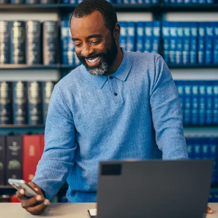 Man in a blue shirt standing in front of library shelves with a laptop and a phone