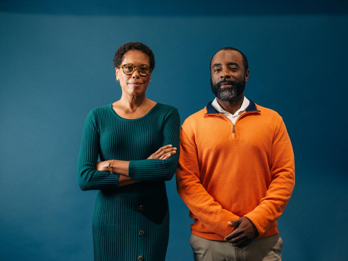 Woman in a dress and man in an orange sweater standing against a blue wall