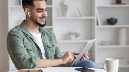 Man in front of a bookcase looking at a tablet and listening on earbuds